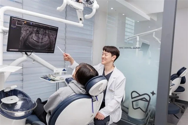 흰 가운을 입은 의료진이 파노라마 엑스레이 화면을 가리키며 진료 의자에 앉은 환자에게 설명하는 모습